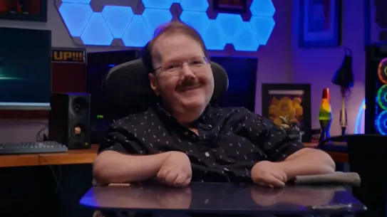 A smiling white man with muscular dystrophy sits at his motorized wheelchair, in front of a monitor gaming setup and blue-lit desk
