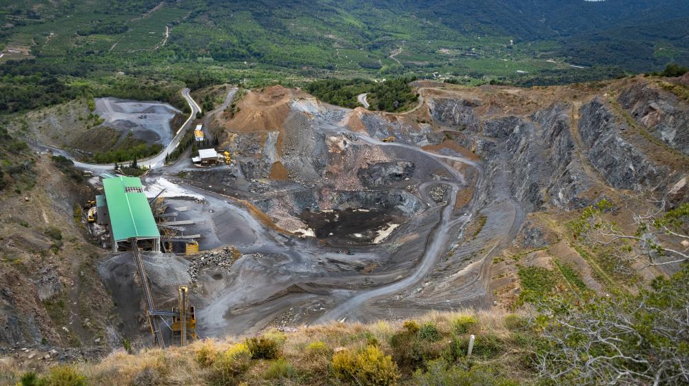 A large mining site with exposed earth, winding dirt roads and industrial structures surrounded by greenery and hills. Credit photo: Envato Elements