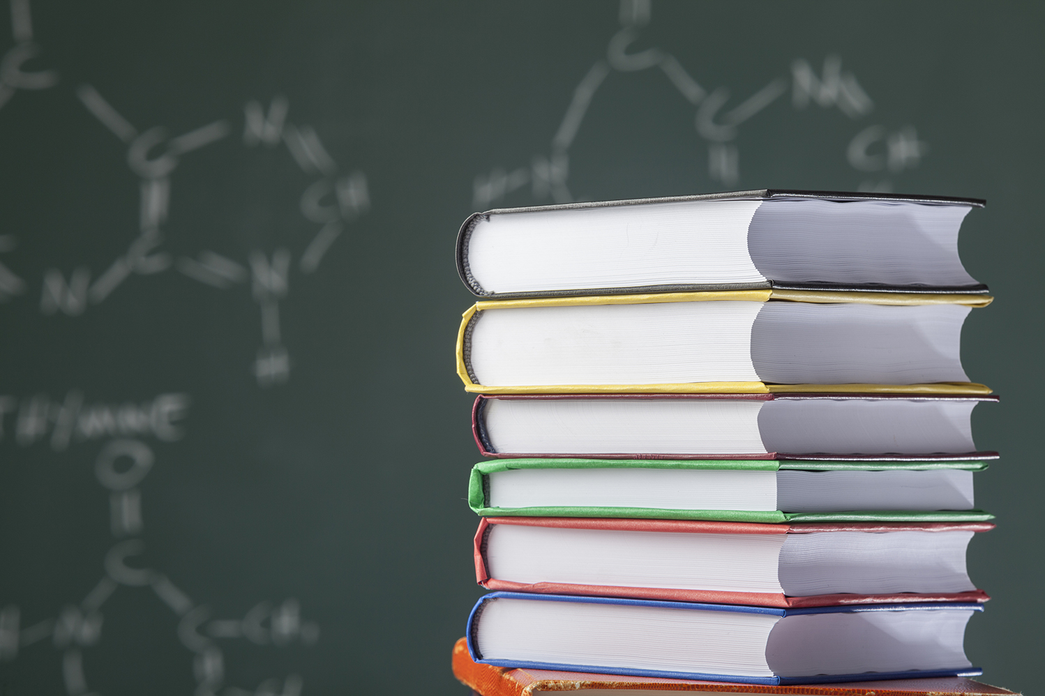 Stack of multicolored books on blackboard with organic molecules written on.The books are placed on the right side of frame.Human DNA formulas are written on board.The focus is on the books and blackboard is blurred.The photo was shot with a full frame DSLR camera in studio lighting.