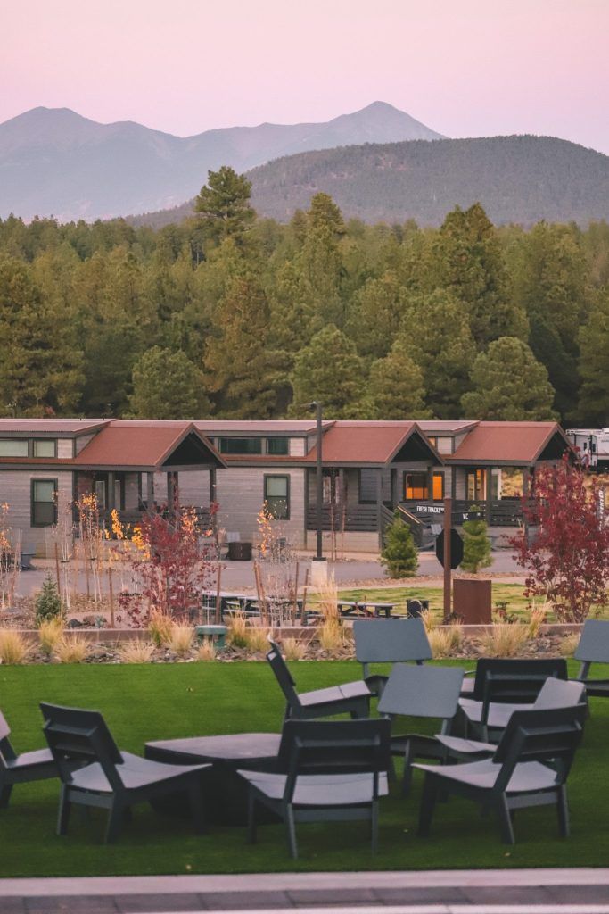 A golden hour mountain range photo with a couple of wooden cabins in the shot. There are shrubs and black seating arrangements at the bottom of the photo.