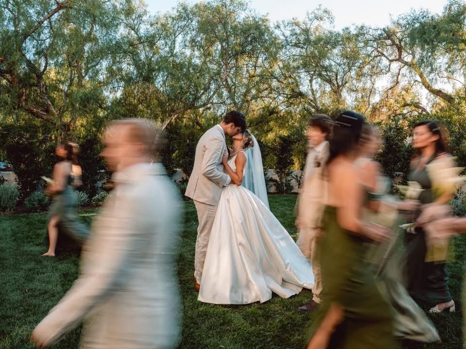 A wide lifestyle shot of a wedding couple sharing a crisp, focused kiss in the center of a grassy lawn, surrounded by the wedding party, who walk quickly past the camera, their bodies stretched into artistic, horizontal motion blurs that frame the scene with a sense of dynamic movement.